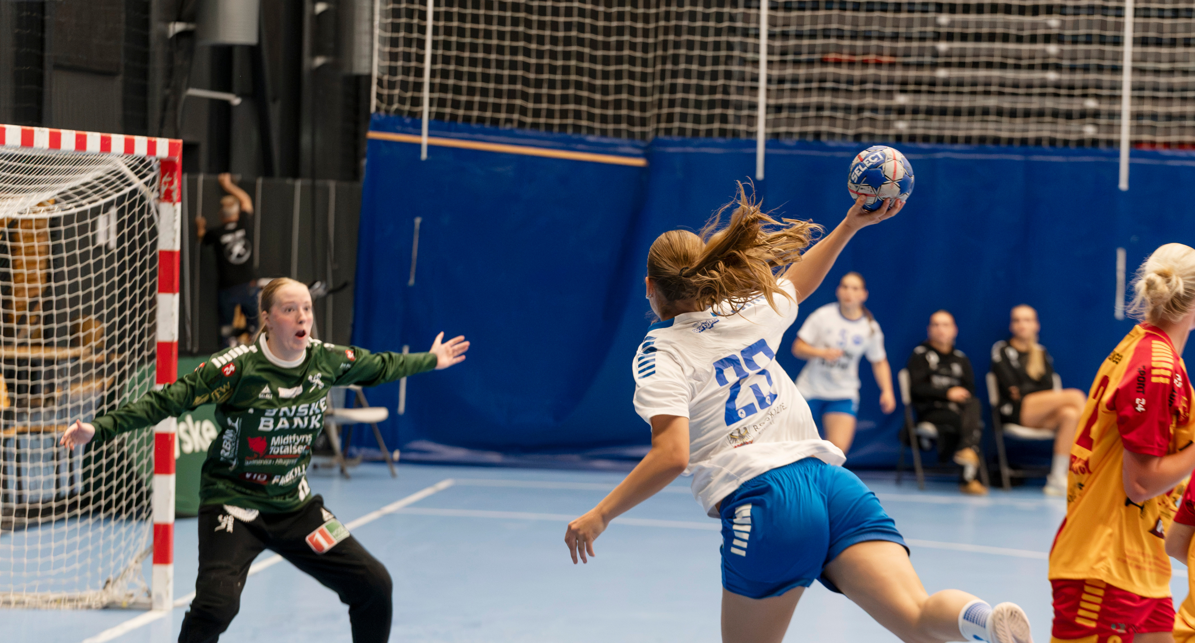 Håndboldspiller i hvid og blå afslutter mod mål, mens målmanden gør sig klar til at redde skuddet. 