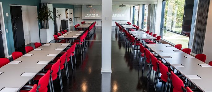 Maglekilde indrettet til møde eller konference med lange bordopstillinger, røde stole og dagslys fra store vinduespartier.