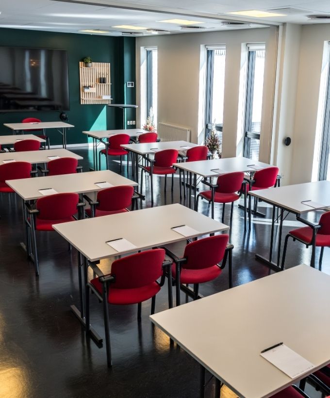 Lovise indrettet med borde og røde stole i skolebordsopstilling samt storskærm og dagslys fra høje vinduer.