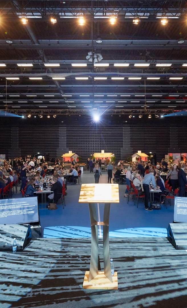 Stor konferencesal med scene, talerstol og deltagere samlet ved borde.