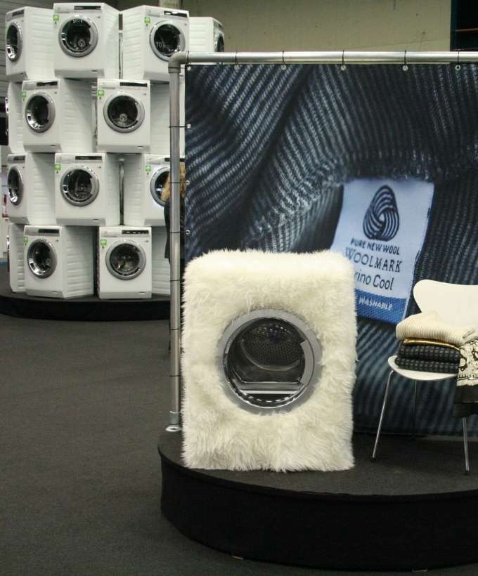 Udstilling af hvidevarer på messe i Roskilde Stand på en messe i Roskilde, hvor hvidevarer præsenteres kreativt med vaskemaskiner stablet i en installation og en udstillet model beklædt med hvidt stof.