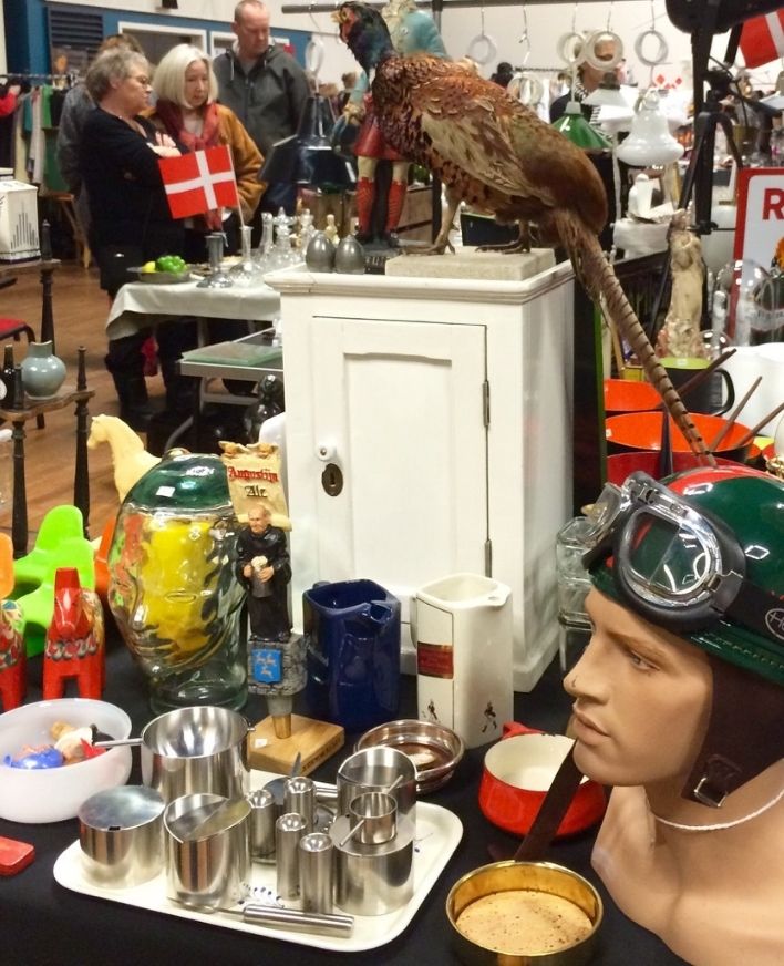 Loppe- og kræmmermarked i Roskilde med retrofund Bord fyldt med loppefund og retro-genstande som porcelæn, metalvarer, en mannequin med pilothjelm og en udstoppet fasan, mens besøgende kigger rundt på markedet.