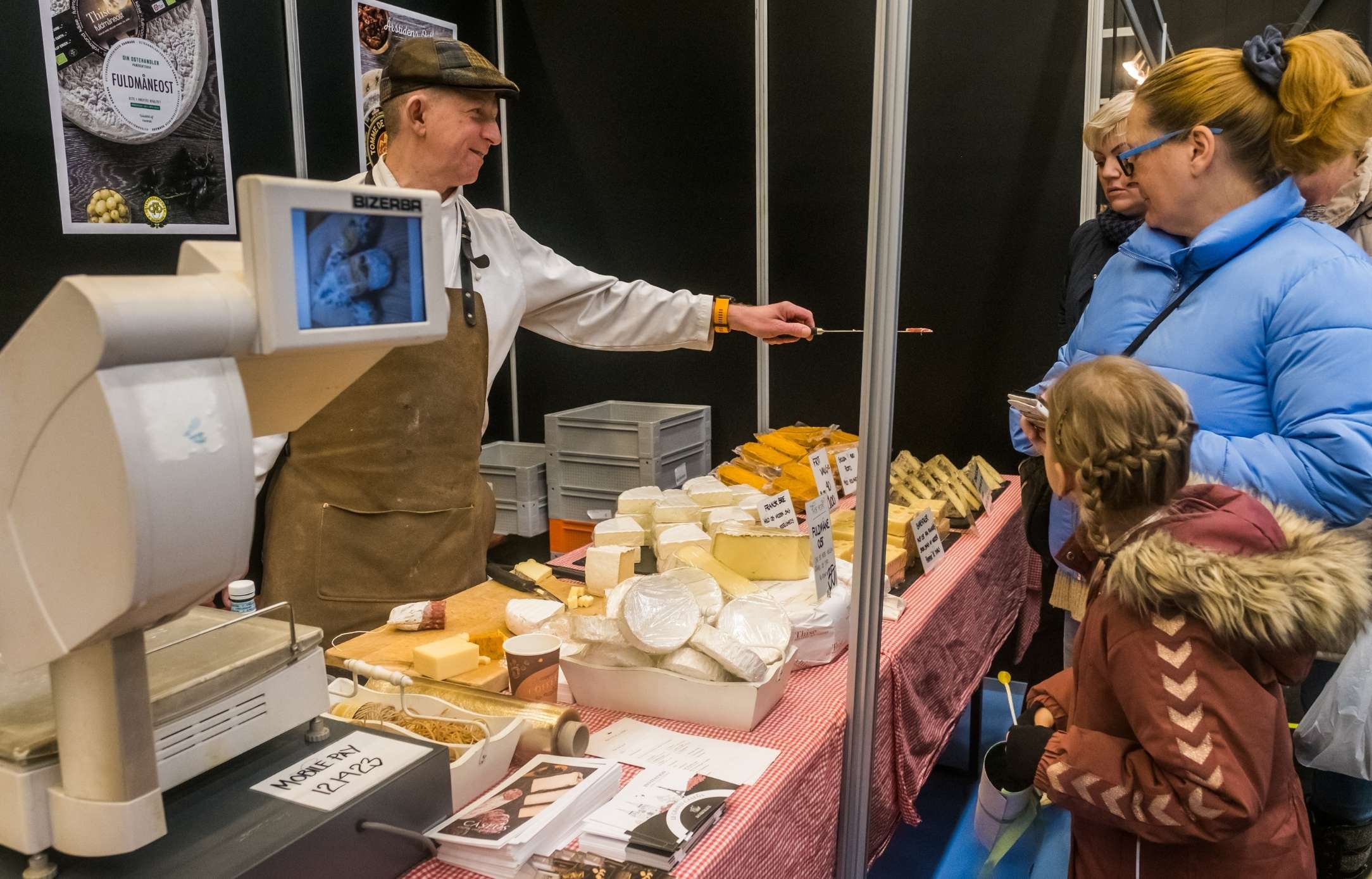 Osteudstiller med smagsprøver på messe i Roskilde Kongres- og Idrætscenter Osteudstiller tilbyder smagsprøver til besøgende på en messe i Roskilde Kongres- og Idrætscenter, hvor lokale delikatesser præsenteres.