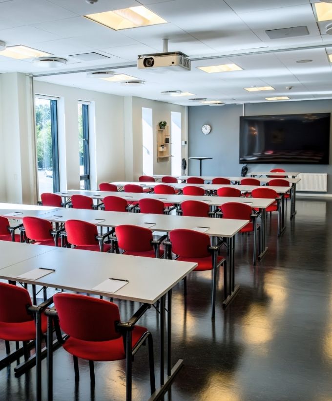 Roar indrettet med borde og røde stole i skolebordsopstilling samt storskærm og projektor.