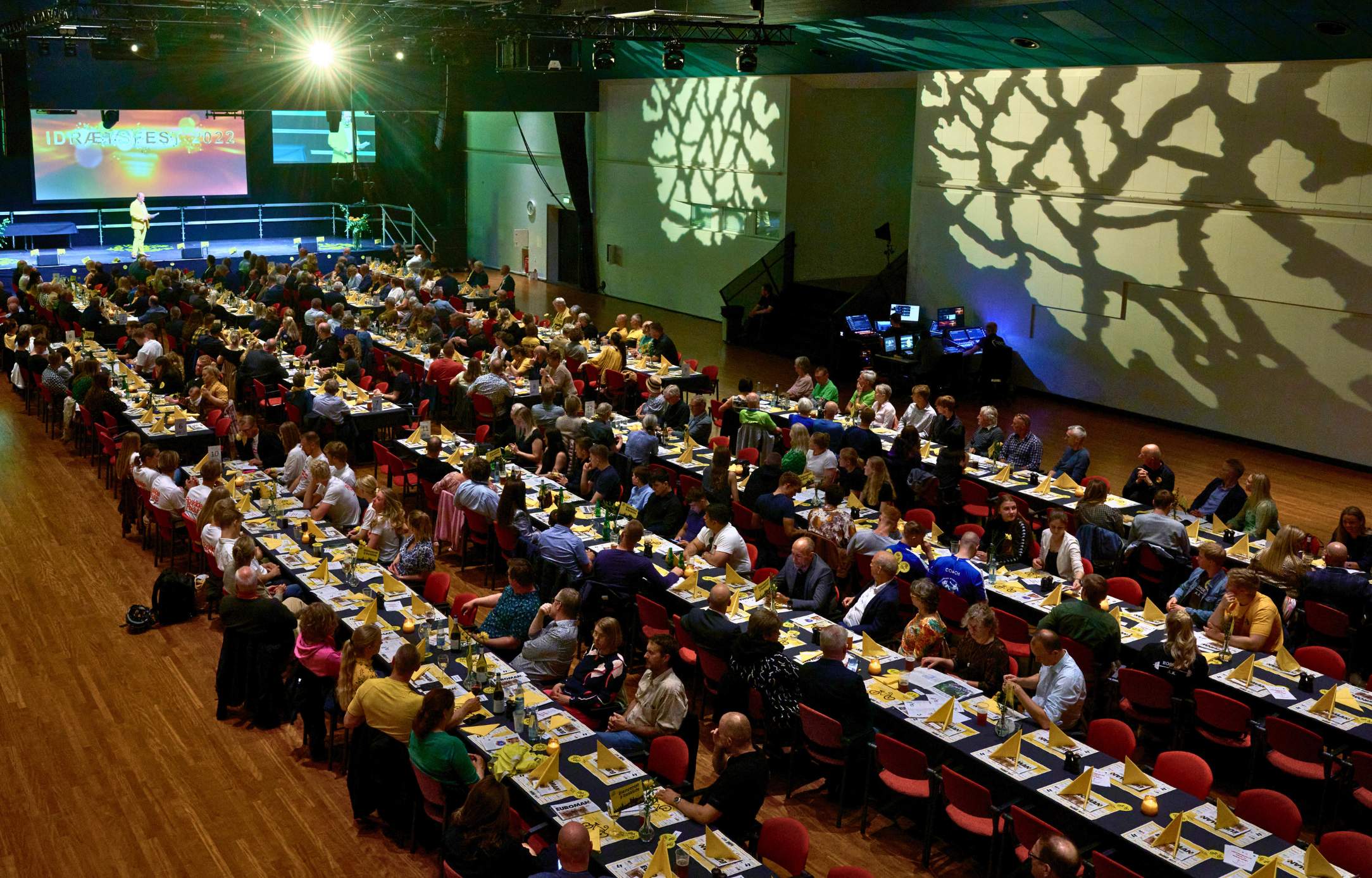 Stort selskab med langborde og scene under en firmafest i Roskilde Kongrescenter (sal A), hvor hundredevis af gæster nyder middag og underholdning.