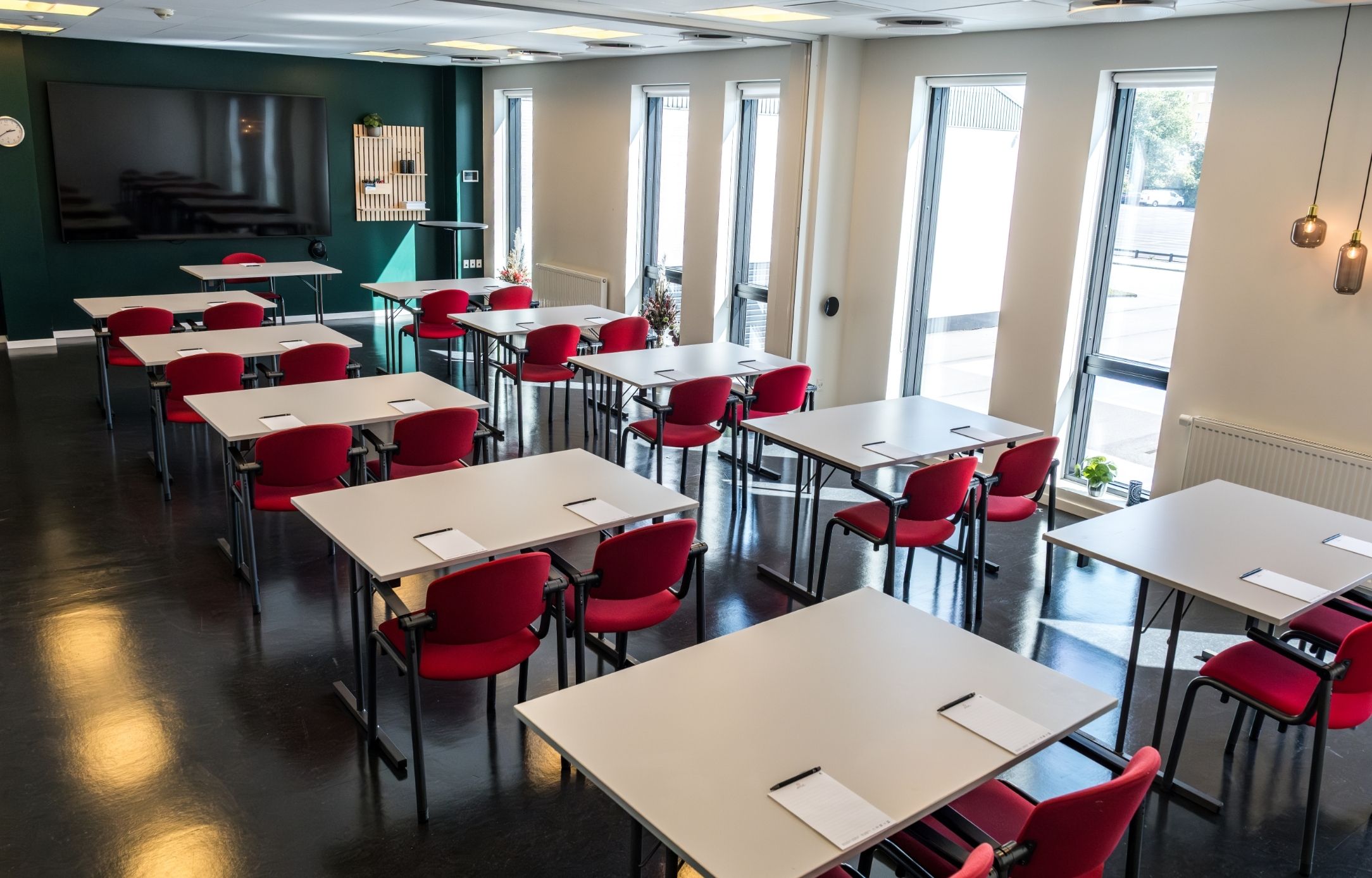 Lovise indrettet med borde og røde stole i skolebordsopstilling samt storskærm og dagslys fra høje vinduer. 