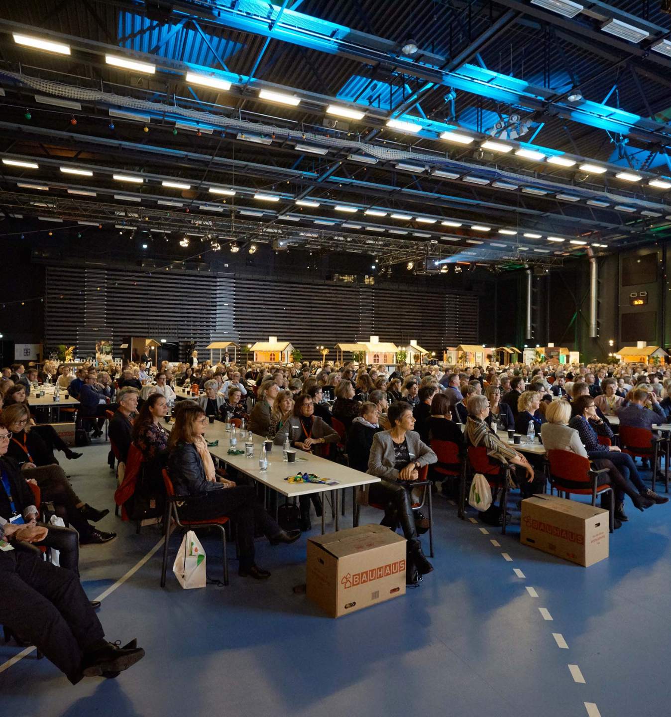 Firmaevent i stor hal med scene, skærme og deltagere ved bordopstilling