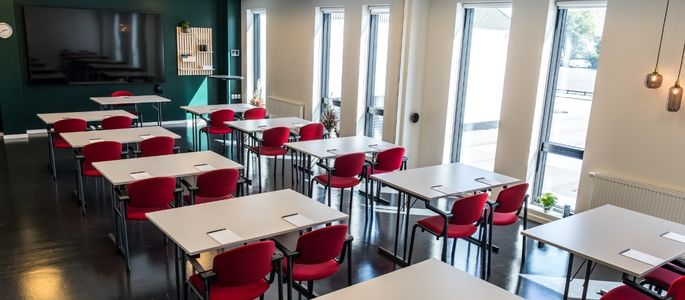  Lovise indrettet med borde og røde stole til møde eller undervisning, storskærm og dagslys fra høje vinduespartier.