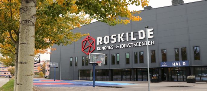 Forpladsen ved Hal D med indgang til hallen, udendørs basketkurve og facade med Roskilde Kongres- og Idrætscenters logo. 