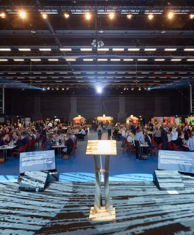Talerstol på en scene med udsigt over deltagere ved langborde i en stor sal.