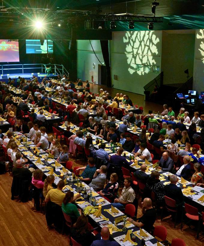 Stort selskab med langborde og scene under en firmafest i Roskilde Kongrescenter (sal A), hvor hundredevis af gæster nyder middag og underholdning.