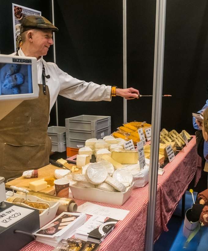 Osteudstiller med smagsprøver på messe i Roskilde Kongres- og Idrætscenter Osteudstiller tilbyder smagsprøver til besøgende på en messe i Roskilde Kongres- og Idrætscenter, hvor lokale delikatesser præsenteres.