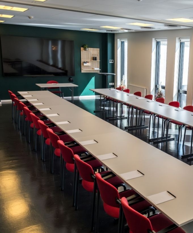 Lovise indrettet med borde og røde stole i hesteskoopstilling foran en stor skærm.