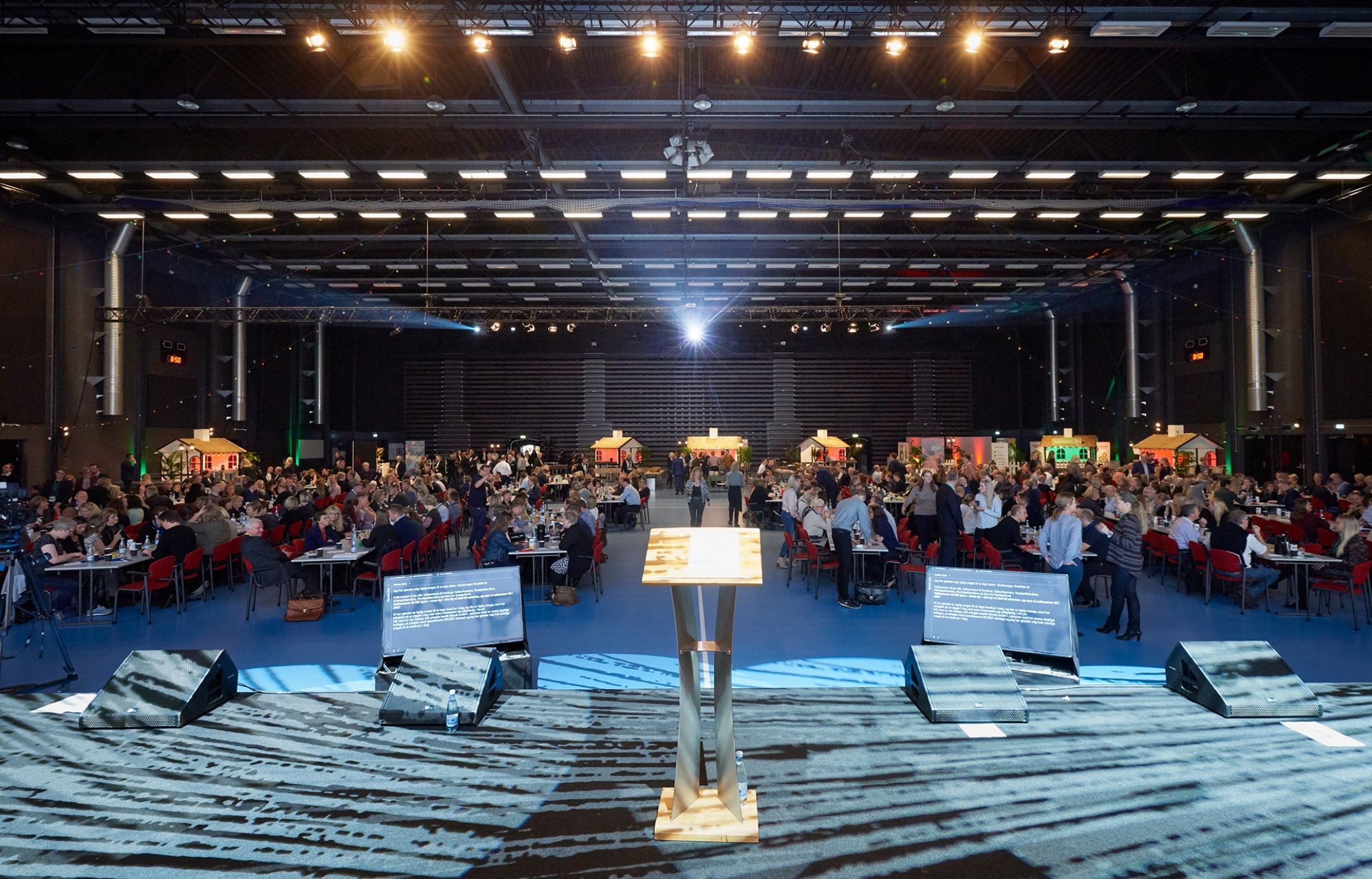 Konferencecenter på Sjælland med plads til store erhvervsarrangementer Stor konferencesal i Roskilde Kongres- og Idrætscenter på Sjælland set fra scenen, med talerstol, lysopsætning og hundredevis af deltagere ved runde borde.
