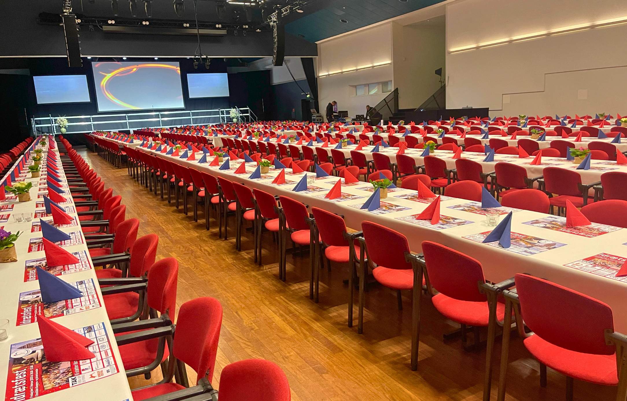 Festlig opdækning med lange borde, røde stole og farvede servietter i Sal A1 med scene.