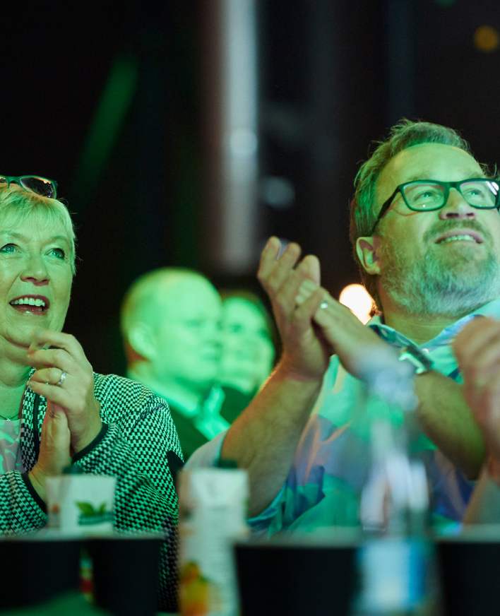 Mand smiler og klapper under en konference i Roskilde Kongres- og Idrætscenter på Sjælland, hvor deltagerne nyder oplæg og fællesskab.