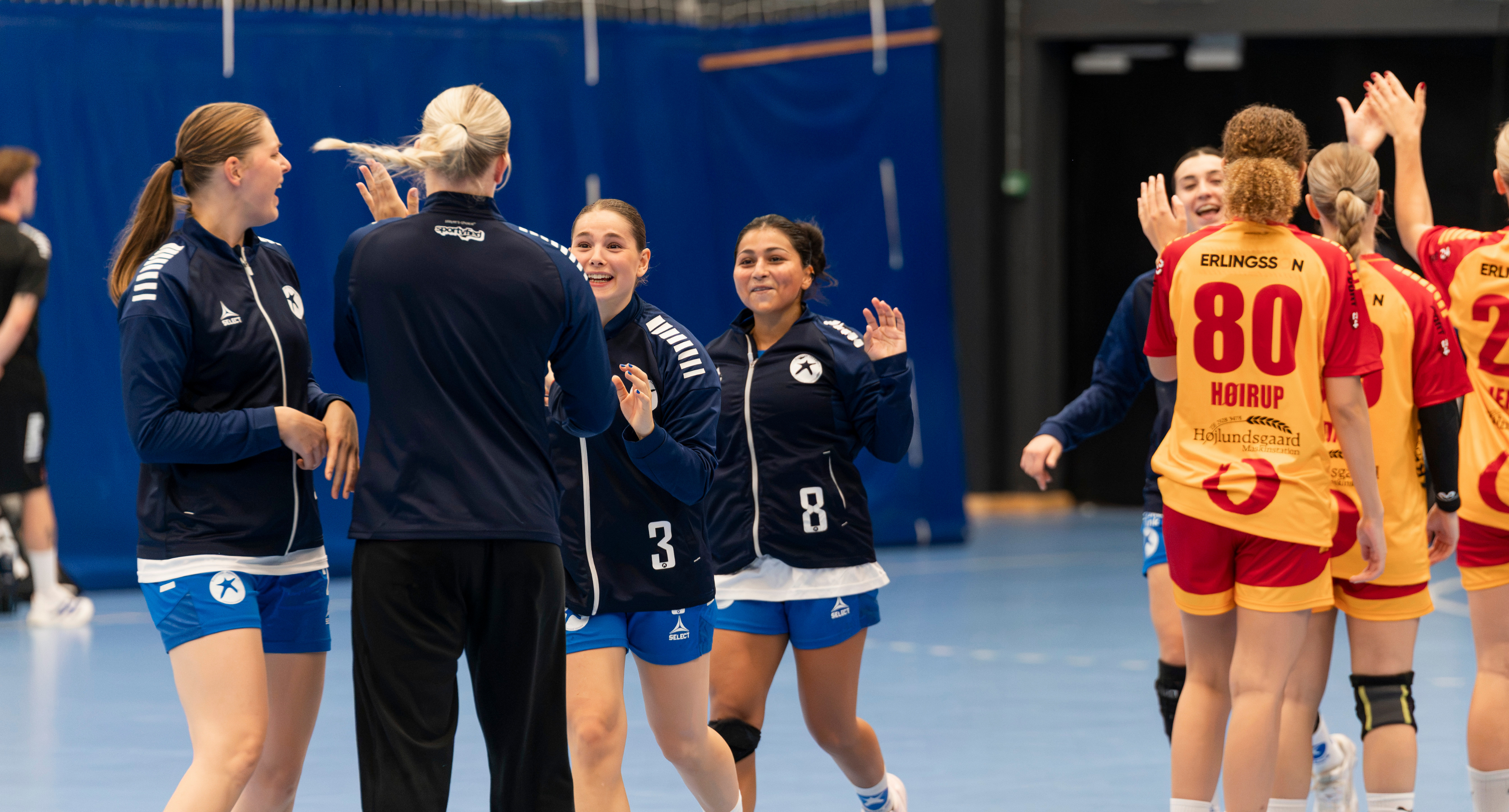 Håndboldspillere i hvid og blå samles på sidelinjen under en kvindehåndboldkamp i Roskilde Kongres- og Idrætscenter.
