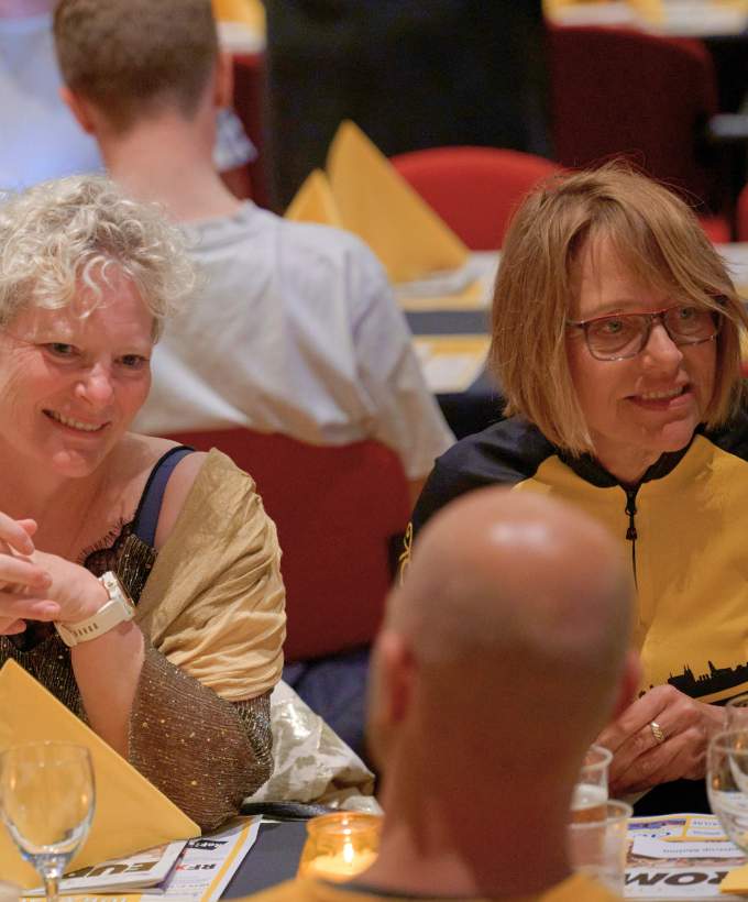 To kvinder smiler og taler sammen ved et festdækket bord under en sommerfest i Roskilde Kongres- og Idrætscenter med hyggelig stemning og farvede servietter.