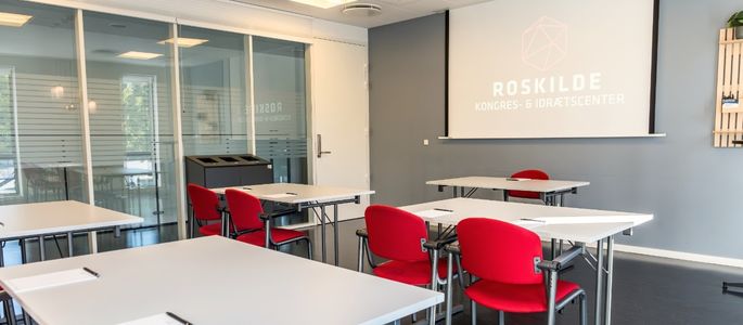Roar indrettet med borde og røde stole i skolebordsopstilling foran lærred med projektor. 