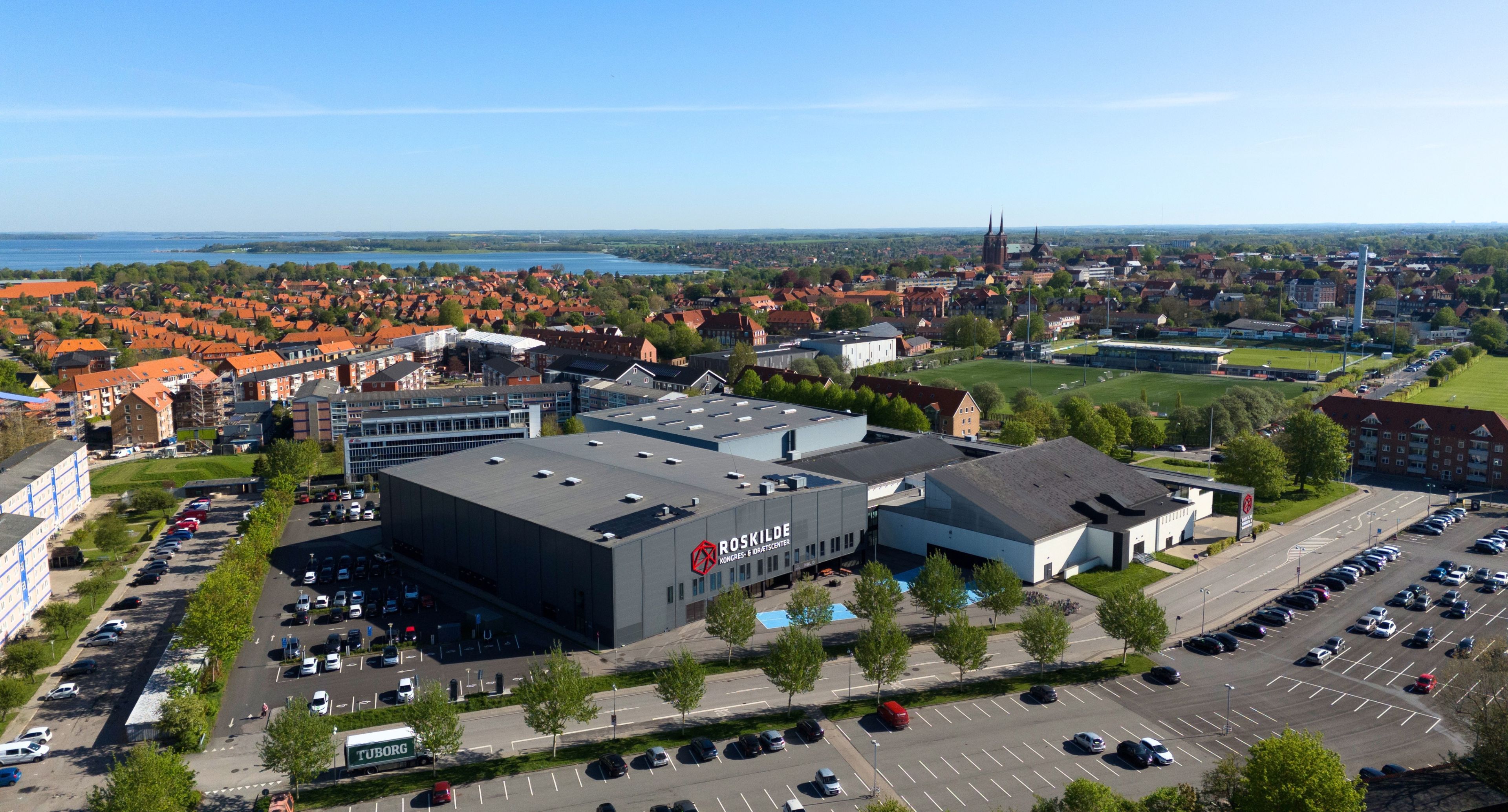 Luftfoto af Roskilde Kongres- og Idrætscenter med parkeringsarealer og Roskilde by i baggrunden.