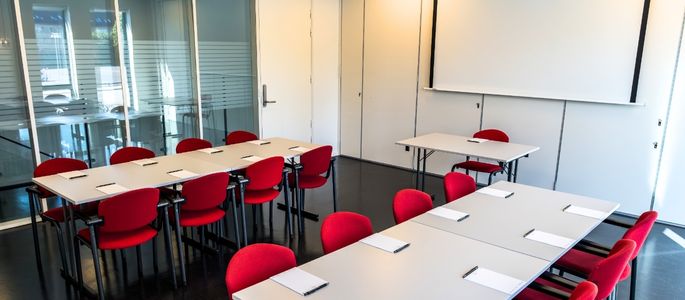 Lovise indrettet med borde og røde stole til møde eller gruppearbejde samt lærred og dagslys fra vinduespartier. 