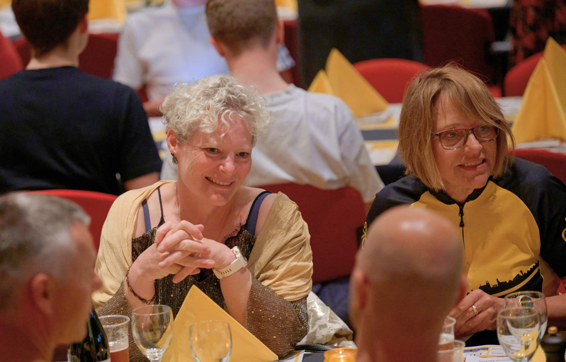 To kvinder smiler og taler sammen ved et festdækket bord under en sommerfest i Roskilde Kongres- og Idrætscenter med hyggelig stemning og farvede servietter.