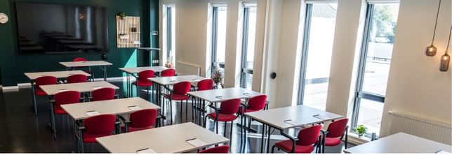Lovise indrettet med borde og røde stole i skolebordsopstilling samt storskærm og dagslys fra høje vinduespartier.