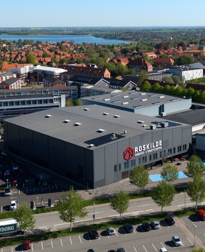 Luftfoto af Roskilde Kongres- og Idrætscenter med parkeringsarealer og Roskilde by i baggrunden.
