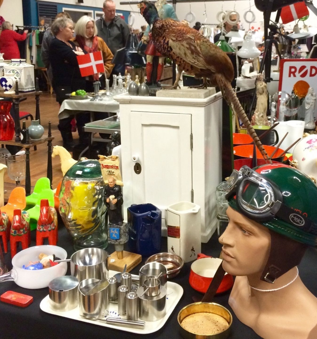 Loppe- og kræmmermarked i Roskilde med retrofund Bord fyldt med loppefund og retro-genstande som porcelæn, metalvarer, en mannequin med pilothjelm og en udstoppet fasan, mens besøgende kigger rundt på markedet.