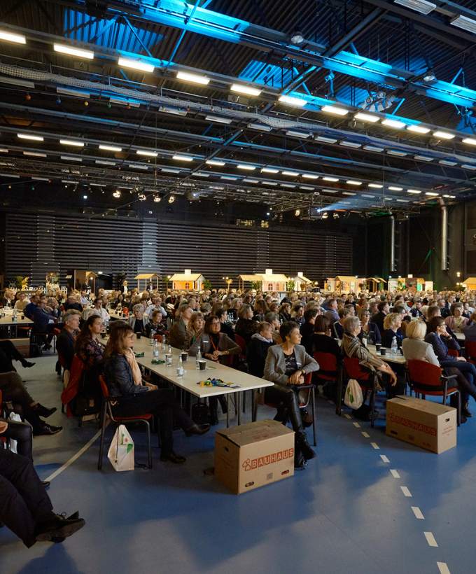Mange konferencedeltagere sidder ved lange borde i en stor hal med scene og teknisk belysning.