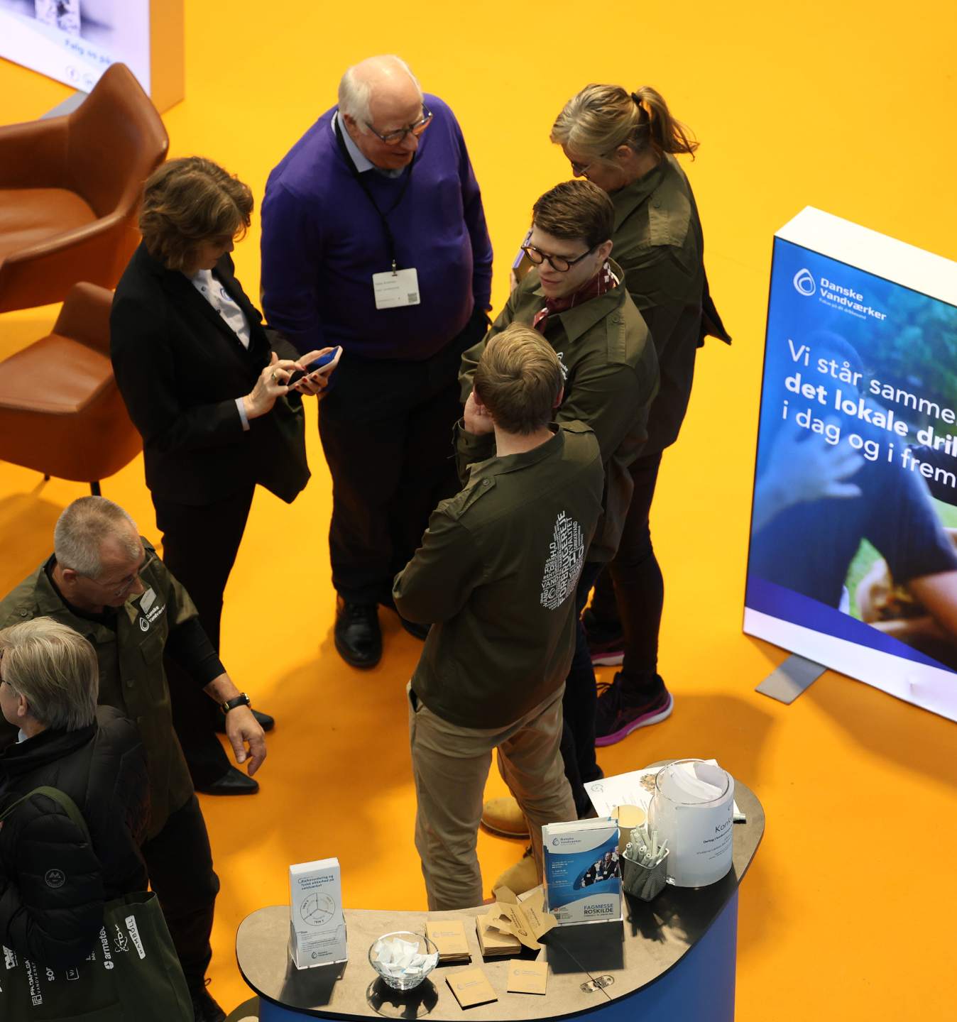 Besøgende i samtale ved en messestand på et gult messegulv, set fra oven.
