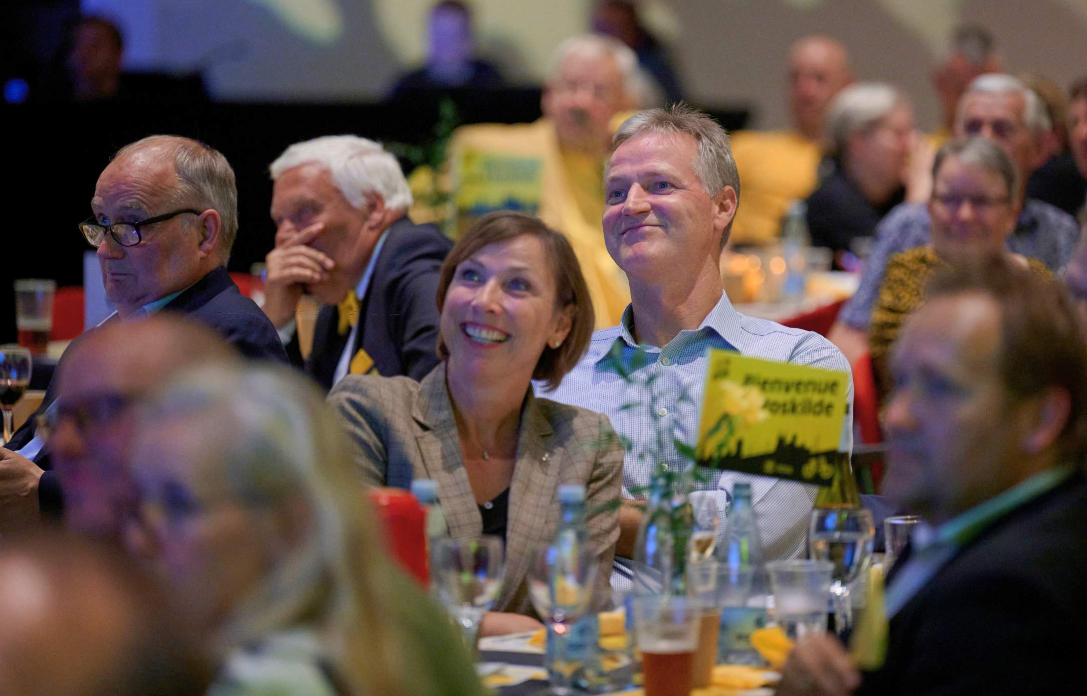 Smilende gæster sidder ved festdækkede borde under et firmaarrangement i Roskilde Kongres- og Idrætscenter med afslappet og hyggelig stemning.